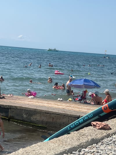 The image depicts a bustling beach scene on a sunny day, likely in Blagoveshchenskaya, Russia, given the presence of a pier and the general atmosphere of a seaside resort. The sea is calm and clear, with numerous people of various ages enjoying the water. Some are swimming, others are floating on inflatable rings, and a few are standing or wading near the shore.
In the foreground, a concrete pier extends into the water, serving as a boundary between the beach and the sea. Several pairs of flip-flops and a child in a pink inflatable ring are visible on or near the pier. A blue beach umbrella provides shade for a group of people sitting on chairs, with a cooler and other beach accessories nearby.
Further out in the sea, several boats are visible. One is a larger passenger boat in the distance, and closer to the shore, smaller boats and watercraft can be observed. Some individuals are engaged in activities like stand-up paddleboarding, as evidenced by a paddleboard in the foreground. The sky is clear and blue, indicating fair weather. The overall impression is one of a lively and enjoyable summer day at the beach.