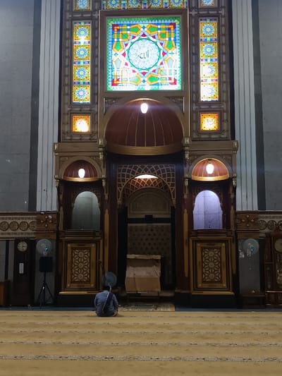 An interior view of Masjid Agung Cianjur (Great Mosque of Cianjur) in Cianjur, Indonesia, shows a single individual seated on a patterned light beige carpet, facing an ornate wooden and stained-glass prayer wall. The person is observed from the rear, wearing a light blue-grey shirt and dark trousers, with a purple-striped sash draped over their left shoulder and back, sitting in a posture indicative of prayer or contemplation.
The dominant feature is the elaborately decorated wall at the front of the prayer hall. It comprises a large central rectangular stained-glass window featuring complex geometric patterns in green, blue, yellow, red, and white, with the Arabic word "Allah" (الله) centrally inscribed. This is flanked by narrower vertical and smaller square stained-glass panels with floral and geometric motifs. Below these, the wall is constructed from dark wood with intricate carvings and golden accents. Two prominent wooden domes with integrated light fixtures illuminate the upper section. Further down, multiple arched alcoves and recessed areas are visible, some also illuminated. The central mihrab niche, appearing dark, is positioned at the base of the ornate structure, with a brown, draped object, possibly a lectern or covered furnishing, positioned in front of it.
On the left side of the scene, near the wall, a blue pedestal fan and a black speaker on a tripod stand are visible, alongside a wall-mounted clock. On the right, another blue pedestal fan and a similar wall clock are present. The room is dimly lit, with focal points of light emanating from within the ornate wall structure and the stained-glass panels. The floor is covered by a light-colored carpet featuring darker patterned stripes extending towards the foreground.