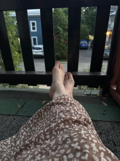 The image shows a first-person perspective from a balcony or porch in McMasterville, Canada. The foreground is dominated by the lower half of a person wearing a brown dress with a white floral pattern, and their bare feet are resting on a dark railing. Beyond the railing, a street scene is visible. A blue house with white-trimmed windows is on the left, and a white car is parked in front of it. Further down the street, a blue car is driving away. Trees and other houses are visible in the background. The lighting suggests it is either dusk or an overcast day.