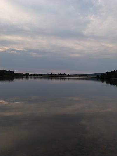 This is a vertical, eye-level shot of a tranquil lake at dusk. The sky is filled with muted, overcast clouds with hints of pink and yellow peeking through the lower portions. The water's surface is calm and reflective, mirroring the sky and the dark, silhouetted treeline that borders the opposite shore. The treeline consists of dense deciduous trees, with some scattered fields and a gentle hill in the background. The overall impression is one of quiet serenity. The location is the lake in Kestřany, Czechia.