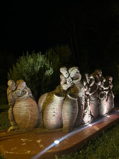This image depicts a striking outdoor sculpture at night, illuminated by artificial lights. The sculpture, rendered in a brick-like material, features several figures grouped around large, vase-like structures. In the foreground, the sculpture rests on a reddish-brown base. The base is lined with small, circular lights that cast beams of light upwards onto the sculpture and the surrounding ground.

The sculpture itself is detailed, with the figures appearing to be engaged in communal activity. The texture of the brick material is evident, suggesting a rough and earthy aesthetic. The lighting creates dramatic shadows and highlights, emphasizing the contours and forms of the artwork.

Behind the sculpture, lush green foliage is visible, also illuminated by lights, creating a vibrant contrast with the dark night sky. The environment appears to be an outdoor setting, likely a garden or public space.

This sculpture is located in Anduze, France, at the Maison de la Céramique, an institution dedicated to ceramics.