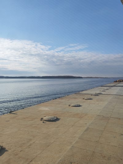 The image displays a wide, calm body of water alongside a large, flat deck, under a bright, partly cloudy sky. The deck, occupying the right side and foreground, appears to be constructed of weathered, light brown metal or concrete panels, showing signs of age and discoloration. Several circular, dome-shaped hatches are evenly spaced along the deck, running parallel to the water's edge, accompanied by smaller mooring cleats.
The water itself is a deep blue-grey, exhibiting gentle ripples and small waves that reflect the daylight. Across the water, a distant shoreline is visible, characterized by a long, dark band of trees that stretches to the horizon. In the far distance on the right, faint yellowish structures hint at industrial or port activity. The sky above is a vibrant light blue, adorned with scattered white stratocumulus clouds and some higher wispy cirrus formations, with sunlight breaking through.
The scene conveys a sense of peaceful tranquility, with no people or specific activities depicted. The lighting suggests mid-morning or afternoon on a clear day, with fair weather conditions. Given the context of Andrukovo, Russia, this is likely a large river or a substantial lake. No text is visible in the image.