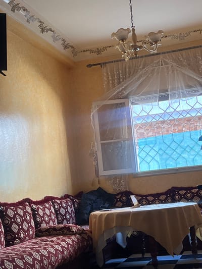 This image depicts the interior of a room, likely a living area or salon, located in Ksar-el-Kebir, Morocco. The walls are finished with a textured, light orange or yellow plaster. A white ceiling features a decorative floral border in a light metallic tone along its perimeter. A three-arm chandelier with cream-colored, scalloped glass shades hangs centrally from the ceiling. A rectangular window, with white frames, is partially open on the right side. Sheer white curtains are present, and a finer white mesh net is draped over the open window portion, resembling a canopy. Beyond the window, an ornate metal grate with a geometric pattern is visible, followed by a light blue element and a brick wall. Below the window, traditional L-shaped Moroccan seating (sedari) extends along the walls. The seating is upholstered in a deep red fabric patterned with white or cream geometric motifs, featuring fringed edges on the cushions. A long, narrow cushion rests in the corner of the seating arrangement. A dark, possibly fabric object, rests on the cushions near the window. In front of the seating, a square wooden table is covered with a light yellow/beige tablecloth layered over a white undercloth. A small, white, rectangular object, possibly a phone stand or small sign, stands upright on the table, with a dark, unidentified object nearby. The floor, partially visible, consists of black and white checkered tiles. A dark, rectangular object, possibly a television screen, is mounted on the wall in the upper left quadrant. No individuals are present in the scene.