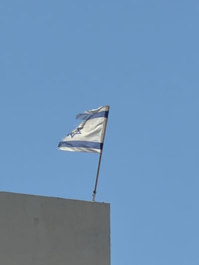 An Israeli flag, torn and tattered, flies against a clear blue sky from a pole extending from atop a building. The flag is white with two horizontal blue stripes and a blue Star of David in the center. The building appears to be a light-colored, flat-topped structure. The scene is captured from a low angle, looking upwards. The image is set in Ashdod, Israel.