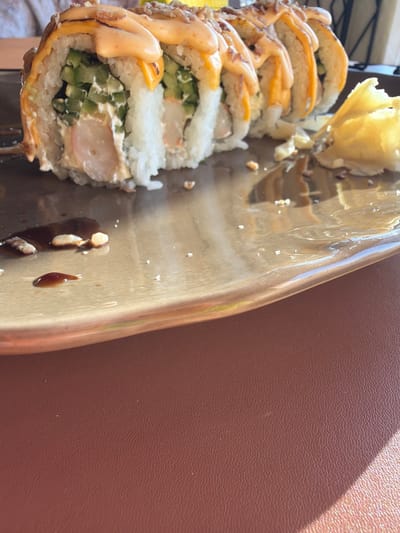 A close-up shot focuses on a plate of sushi, possibly in Khotianivka, Ukraine. The sushi rolls are topped with a creamy orange sauce and crispy bits. To the right, a dollop of yellow pickled ginger sits on the plate. The plate itself has a metallic sheen, reflecting the light and the sushi. Scattered on the plate are small remnants of soy sauce and white rice. The lower portion of the image reveals a textured brown surface, likely a table, with a distinct shadow cast across it from the sunlight. No individuals are clearly visible in the frame, though a hint of clothing is present in the upper left corner.