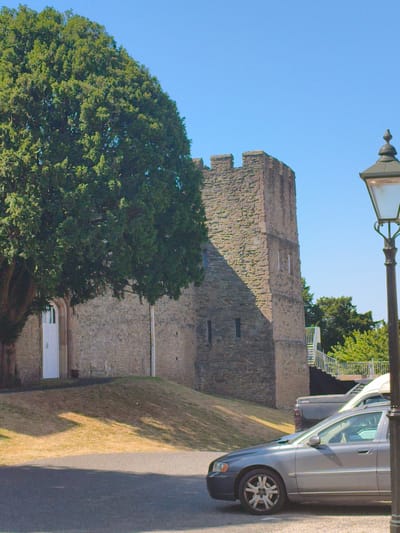 A grey sedan is parked in the foreground on a paved road. Behind the sedan, a grassy slope rises towards a stone building that appears to be part of a castle. The building has a tall, square tower with crenellations and small windows. A large, leafy tree with dark green foliage is situated to the left of the stone building. A white door is visible on the side of the building. In the background, there are more trees and a clear blue sky. A lamppost with a black base and green lantern is positioned on the right side of the image. Another vehicle, a dark colored pickup truck, is parked behind the sedan. The image appears to be taken on a sunny day. The context of Ludlow, United Kingdom is not explicitly visible in the image itself but may be known from metadata or external information.