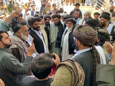 The image shows a group of men gathered in an open-air space in the city of Zaker-E Sharif, Afghanistan.  The men are mostly wearing traditional clothing and many are wearing turbans.  The image appears to have been taken in a courtyard or plaza near a mosque as there are people in the background exiting the mosque. There is an older man at the center of the image. He is surrounded by men who are all looking at him and speaking. The man is wearing a white shirt and black vest, and he appears to be in a position of authority. Some men are holding cell phones. The men appear to be engaging in a conversation and there is a lot of interaction happening. The environment is open air, likely a courtyard or a public space. The architecture in the background suggests a traditional Afghan building.