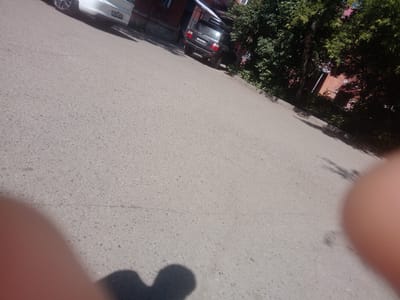 The image is a low-angle shot of an asphalt street in Oskemen, Kazakhstan, on a sunny day. A white car with a license plate ending in "02" is parked on the left side of the frame. Further down the street, a dark-colored SUV with a license plate reading "703 ANZ 16" is parked. Lush green trees and a brick building are visible in the background. The foreground is partially obscured by a blurred human finger and shadow. The street appears to be a residential or semi-commercial area.