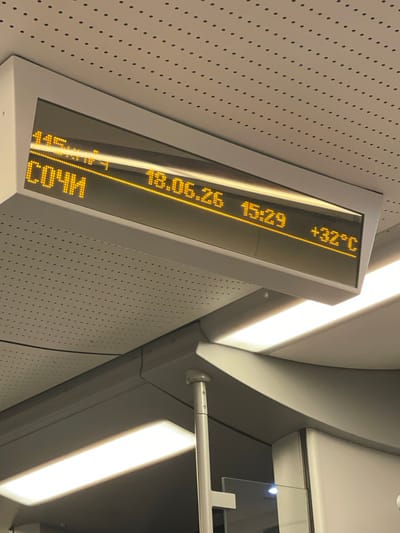 This image captures a digital display inside what appears to be a train or a similar public transportation setting in Monastyr, Russia. The display is oriented vertically and shows information in bright yellow LED characters against a dark background.
The visible text on the display includes a date and time, "18.06.25 15:29," and a location, "СОЧИ" (Sochi). It also shows a temperature reading, "+32°C". The setting is dimly lit, with bright overhead lights creating strong reflections on the display and surrounding surfaces. The wall behind the display is textured with a pattern of small, regular dots. The overall impression is of a functional, modern interior, likely within a travel context. There are no people visible in the image. The time of day is mid-afternoon, and the temperature indicates warm weather.