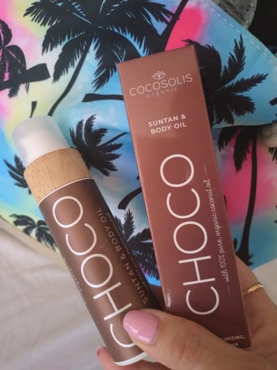 The image shows a close-up of two products from the brand Cocosolis, a suntan and body oil. One is a rectangular box, and the other is a cylindrical bottle with a wooden cap. Both products feature the branding "CHOCO" in large white letters against a brown background, along with smaller text indicating "SUNTAN & BODY OIL" and "with 100% pure, organic coconut oil." A woman's hand with pink nail polish and a gold ring is visible, holding the cylindrical bottle. The products are placed on a colorful fabric with a tropical palm tree print, suggesting a beach or vacation setting. The lighting appears to be natural, possibly from daylight. The overall impression is one of summer and self-care. There is no specific activity or event taking place beyond the presentation of the products.