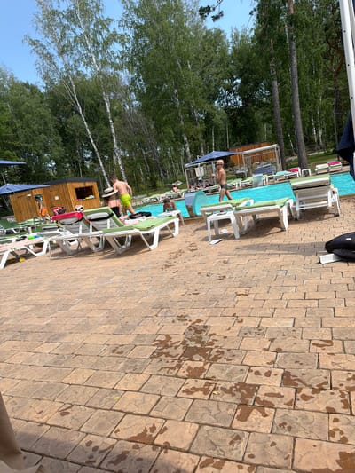 The image shows a poolside scene with several people and lounge chairs. In the foreground, a person in green shorts is standing near a lounge chair, and another person wearing a hat is lying on a lounge chair. Further back, there are more lounge chairs and a swimming pool. Beyond the pool, a line of trees forms a backdrop. The ground around the pool is paved with large, irregularly shaped stones that appear wet in places, suggesting recent activity or a spill. The lighting indicates it is daytime, with sunlight casting shadows. The setting appears to be a resort or a public pool area.