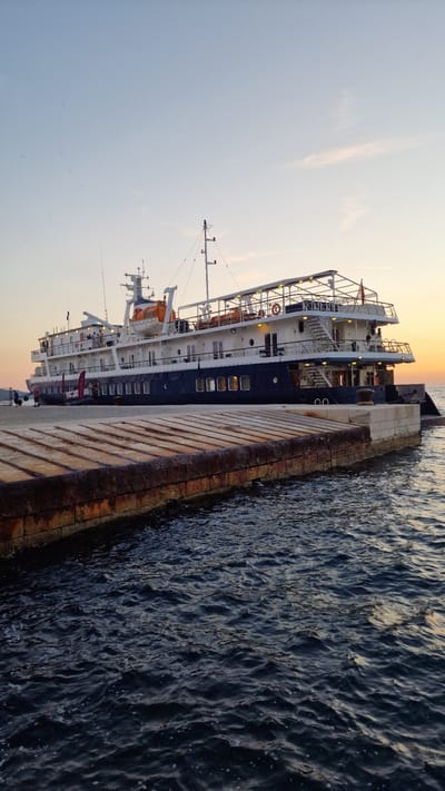 This media file captures a serene scene at sunset in Zadar, Croatia. The primary subject is a large, multi-decked vessel, likely a ferry or cruise ship, docked alongside a stone pier. The ship is predominantly dark blue with white upper decks, featuring multiple windows and railings. Life rafts are visible, adding to the nautical theme.

The activity taking place appears to be the ship at rest, docked at the harbor. There are no prominent activities of people visible, suggesting a quiet moment. The time of day is clearly sunset, with the sky displaying a gradient of soft oranges and yellows fading into a pale blue, indicating the end of the day. The water is dark and appears slightly rippled, reflecting the ambient light. The pier is made of weathered stone, showing signs of age and exposure to the elements, with a rusty edge suggesting it's a well-used structure.

The location cues, specifically the mention of Zadar, Croatia, help contextualize the image. The overall mood conveyed is peaceful and tranquil, characteristic of a sunset by the sea. No visible text is discernable in the image.