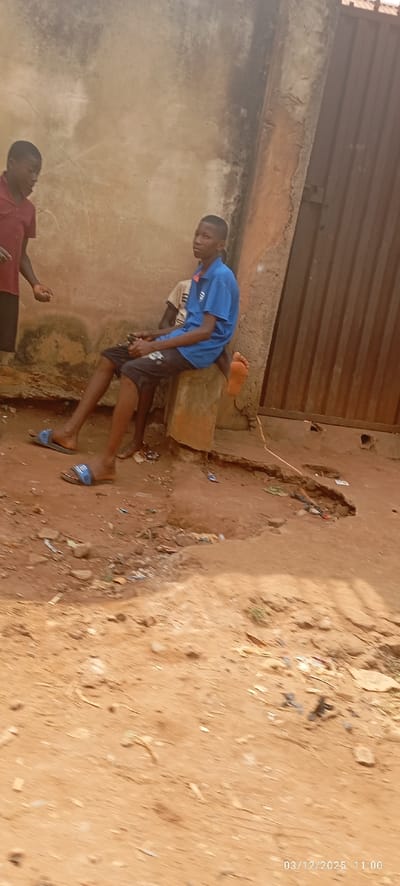 The image depicts an outdoor scene in Abagana, Nigeria, at approximately 11:00 AM on March 12, 2025. Three young males are positioned on uneven reddish-brown dirt ground. The background features a textured, light-brown plastered wall on the left, transitioning into a darker concrete section and a brown, vertical-paneled metallic gate with a visible latch mechanism on the right.
A young male, wearing a red short-sleeved collared shirt and dark trousers, stands on the left side of the frame, facing right with his left hand slightly open. In the center, a second young male, attired in a blue short-sleeved collared shirt with a light placket and dark shorts, is seated on a low, light-colored concrete block. He wears blue slide sandals on his left foot, while his right foot is bare and elevated. He is looking forward, directly at the viewer. Partially obscured behind him and also seated, a third young male is visible, wearing a light-colored short-sleeved shirt and dark shorts, with bare feet. The ground exhibits scattered small pieces of debris and litter.