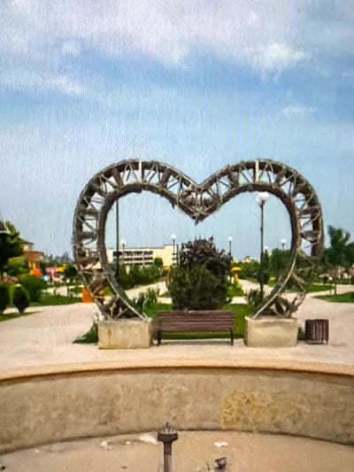 This is a vertical, slightly blurry photograph taken outdoors. The main subject is a large, open heart-shaped sculpture made of metal beams. The sculpture is positioned in the foreground, dominating the left side of the frame. Behind the sculpture, a pathway or walkway is visible, lined with green grass and some trees. There are also benches and what appears to be a street lamp within the sculpture's opening. Further in the background, there are hints of buildings and more greenery, suggesting an urban park or public space. The sky is visible at the top of the image, appearing cloudy with patches of blue. The overall lighting suggests it might be daytime, possibly late afternoon, with natural light. There is no discernible text in the image. The scene evokes a sense of public art and a pleasant outdoor environment. The sculpture's design is intricate, with a network of supporting beams creating a textured effect.