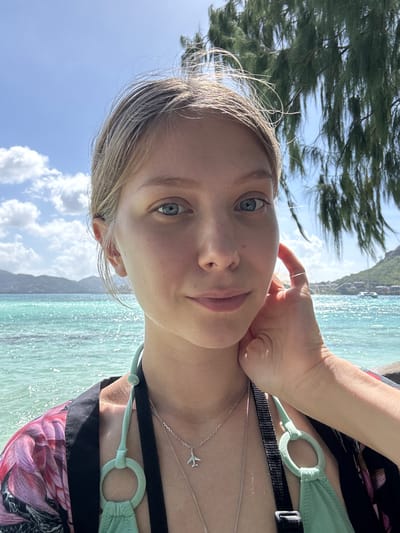 A selfie of a young Caucasian woman with blonde hair and blue eyes is taken from the chest up. She is outdoors, with the ocean and a tree in the background. The ocean is a bright turquoise color and appears to be calm, with small waves. A lush green tree with long, thin leaves hangs over the woman's right shoulder. The sky is a clear blue with a few white clouds. The woman is wearing a light green bikini top and a patterned kimono. She has a silver airplane pendant necklace on. She is touching her face with her right hand. The location is likely a beach or coastal area in Victoria, Seychelles, given the tropical vegetation and clear blue water.