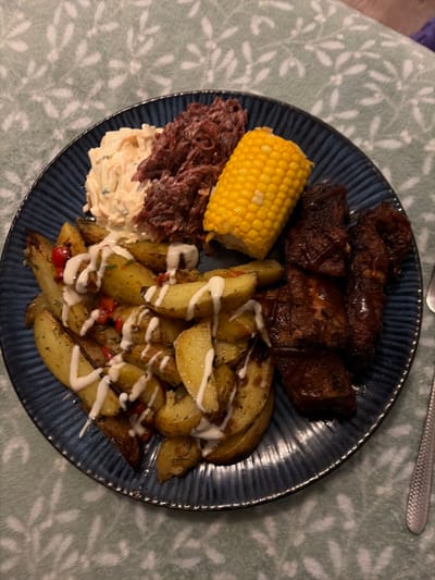 The image displays a single serving of a prepared meal on a dark blue, ridged circular plate, situated on a light green tablecloth with a subtle white leaf pattern. The plate holds multiple food items: roasted potato wedges, seasoned with herbs and red flakes, are piled on the lower-left and drizzled with a white creamy sauce. Adjacent to them on the upper-left, a portion of creamy coleslaw is present next to a mound of dark red, shredded food, possibly red cabbage slaw or pulled meat. Centered at the top of the plate is a half-cob of yellow corn. On the right side, several pieces of dark, glazed meat, consistent with barbecue ribs or chicken wings, are arranged. A silver-colored fork with a textured handle is partially visible along the right edge of the frame. The scene is consistent with an indoor dining setting, specified as being in New Forest, United Kingdom, though the image itself contains no explicit geographical markers. No individuals are present in the image.