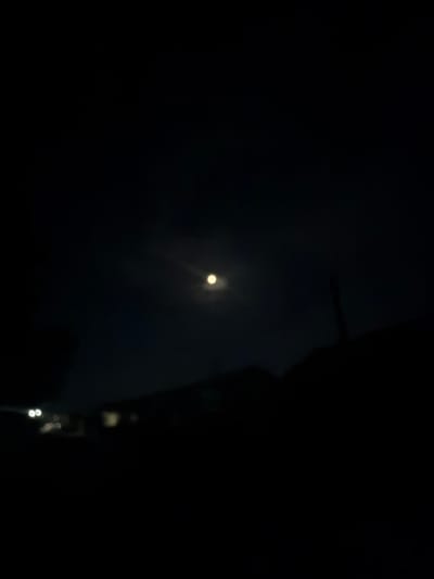 The image captures a dark night scene with a bright moon in the upper center of the frame. The moon is slightly blurred and has a radiant glow. Below the moon, a faint silhouette of trees or structures can be seen against the dark sky. In the lower portion of the image, there are blurry lights that suggest the presence of buildings or streetlights in the distance. The overall impression is of a quiet night, possibly in a rural or suburban setting within the city of Yangiiul, Uzbekistan.