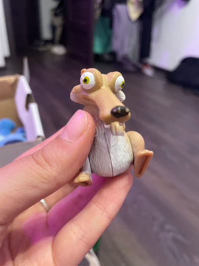 A close-up, eye-level shot shows a plastic toy figure of the character Scrat from the movie "Ice Age." The figure is held by a person's hand in the foreground. Scrat is depicted with its characteristic large eyes, long snout, and visible teeth. The toy is light brown and grey, with some visible seams indicating it is made of multiple plastic pieces. The background is out of focus but appears to be an indoor setting with a wooden floor and some blurred furniture or structures. The image appears to have been taken indoors, possibly in a residential setting. There is no indication of the location being Vaganovo Massiv, Russia.