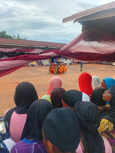This image depicts an outdoor event or gathering in Ogbogbo, Nigeria. The scene is observed from behind a group of spectators in the foreground, looking towards a central activity area.
In the immediate foreground, the backs and profiles of approximately ten individuals, predominantly females, are visible. Most wear head coverings in various colors including black, pink, red, light blue, white, and a lighter green. Specific garment details include a pink shirt, a purple and blue striped shirt, and a yellow, orange, and pink patterned fabric on the lower right. One individual's braided hair is visible beneath a dark head covering.
The midground features an open, reddish-brown earthen ground area. Centered within this area, approximately four to five individuals, appearing to be children or young adolescents, are positioned in brightly colored, elaborately patterned costumes, predominantly yellow, orange, and red with black accents. To their right, a single adult male stands, dressed in a light blue shirt and green trousers. Along the sides of this central area, several white rectangular tables with yellow tablecloths are arranged, accompanied by chairs. Numerous individuals are seated at these tables, observing the central activity.
Spanning the upper midground and separating the foreground spectators from the background, lengths of sheer fabric in maroon and lighter pink are draped horizontally, creating an overhead canopy or decorative element. These fabrics are loosely hung and show some billow.
In the background, a long, single-story building with a reddish-brown, rusted corrugated metal roof and white eaves extends across the frame. Green foliage and trees are visible above and to the left of this structure. The sky is overcast with diffuse cloud cover.
