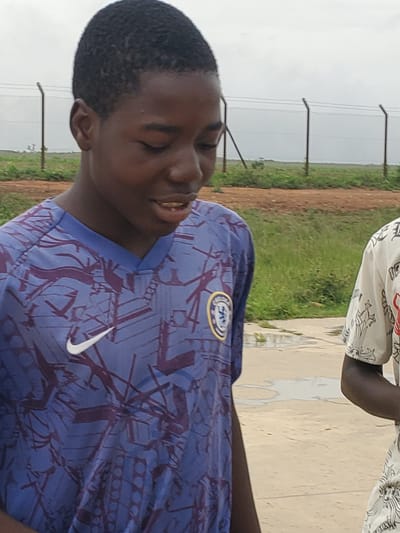 The image shows a close-up of a young Black person wearing a blue t-shirt with a purple abstract pattern and a white Nike logo. The person has dark, short hair and is looking to the right with their eyes closed, a slight smile on their face. The t-shirt has a crest on the left chest, possibly a sports team logo.
In the background, another person is partially visible wearing a white shirt with a black and red pattern. The setting appears to be outdoors, with a fence and grassy area behind the subjects. The ground in front of them looks like a dirt path or road. The sky is overcast, suggesting a cloudy day. The overall scene gives the impression of a casual outdoor gathering or activity. The location context is Pankshin, Nigeria.