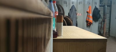 The image shows a close-up, low-angle view of a wooden desk with several items on it. To the left, a portion of a wooden cabinet or drawer is visible, its surface showing vertical wood grain. On the desk, in the center of the frame, is a cylindrical object, possibly a cigarette butt or a small container, with a brown top and a speckled white body. Behind it, two plastic bottles are lined up: one is a blue bottle with a green cap, and the other is a clear bottle with a red cap, showing the Coca-Cola logo. To the right of the bottles, a dark brown leather bag is partially visible. In the background, a row of metal lockers is lined up against a wall. Several items are hanging on or near the lockers: a grey jacket hangs on a hook, an orange high-visibility vest with reflective stripes is on another hook, and a black backpack is also hung up. The environment appears to be a locker room or a changing area, possibly in an industrial setting or a sports facility, located in Salzgitter, Germany. The lighting is dim, creating shadows and highlighting the textures of the objects on the desk.