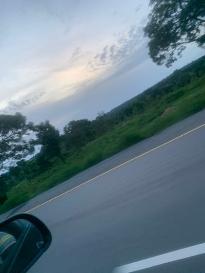 The image depicts a road in Gudi, Nigeria, with lush green vegetation lining the right side of the road, and a cloudy sky above. The perspective suggests the photo was taken from a vehicle, possibly a car, with the side mirror visible on the left. The road itself is asphalt, with a yellow line marking the lane. Trees are scattered along the roadside, and some are blurred due to motion. The overall scene conveys a sense of travel through a rural or semi-rural area.