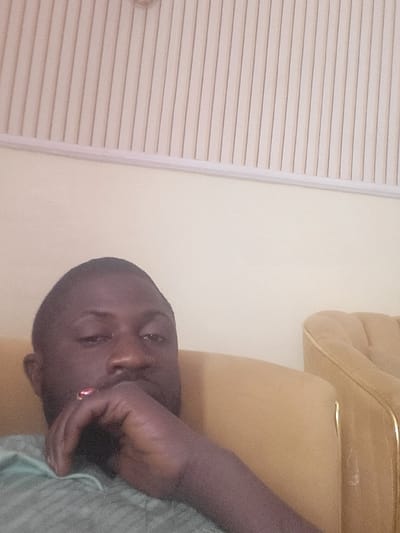 This is an indoor scene located in the city of Babawa, Nigeria. An adult male with dark skin is depicted in a medium close-up, occupying the lower left and center portions of the frame. He is seated on what appears to be a tan or gold upholstered sofa, visible on the right side and beneath him. His right hand is raised, with his chin resting on his knuckles, partially obscuring his mouth. A red ring is visible on his right pinky finger. He is wearing a light green, possibly patterned, shirt. His gaze is directed towards the camera. The background consists of a plain light-colored wall above the sofa, topped by a ceiling featuring light-colored vertical paneling. The lighting suggests an interior environment.