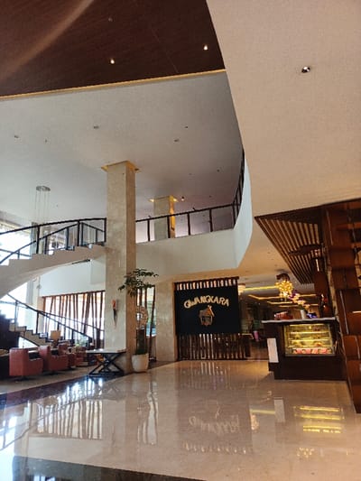 A spacious hotel lobby features polished marble floors reflecting the light. A staircase with black railings curves upwards. A tall marble column anchors the space, adjacent to a seating area with red chairs. The "GIWANGKARA" restaurant entrance is visible with a dark curtain and wooden dividers. A pastry display case stands nearby, illuminated by overhead lights. The ceiling is high, with a mix of white and dark wood panels.