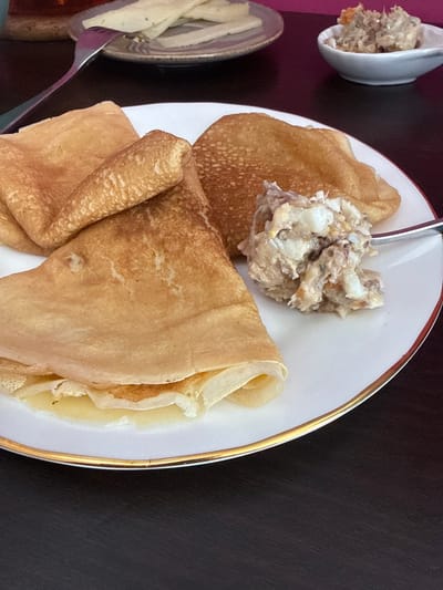 A close-up, overhead shot shows a plate of crepes and a small bowl of what appears to be a fish or seafood salad. The crepes are golden brown and folded, with some appearing slightly crisp. The salad is a creamy mixture with visible chunks of fish or seafood and possibly herbs. A fork rests on a plate in the background, with thin slices of what appears to be cheese or some other food item. The scene is set on a dark, textured surface. The overall impression is a delicious, home-style meal, perhaps for breakfast or brunch. The image was taken in Almada, Portugal.