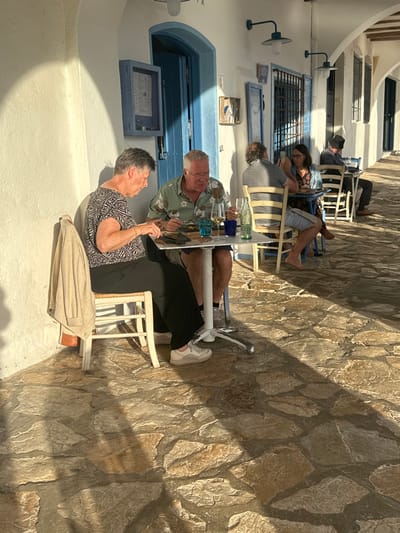 The image displays an outdoor dining area within an arched white colonnade in Palafrugell, Spain. The ground is paved with irregular, light-toned flagstones, with direct sunlight illuminating the left side and casting distinct diagonal shadows across the pavement. The architectural elements consist of white stucco walls, blue painted wooden doors, window frames with grilles, and wall-mounted blue light fixtures. A blue, patterned box-like object is mounted on the wall above the foreground blue door.
In the foreground, an older woman with short grey hair, wearing a patterned short-sleeved top and dark trousers, sits on a white wooden chair. A beige cardigan is draped over the chair's back. She holds cutlery over a plate on a white square table. Opposite her, an older man with short grey hair and glasses, wearing a patterned green short-sleeved shirt and light shorts, is also seated at the table, eating from a plate. The table holds additional items including a wine glass, a blue tumbler, and a clear glass bottle.
In the mid-ground, a second white square table is occupied by a man in a grey shirt and a woman holding a smartphone. The woman is barefoot. Further down the colonnade, a third table shows a man in a dark hat seated with his back partially to the viewer. The background features more white walls and blue accents, illuminated by direct sunlight.
