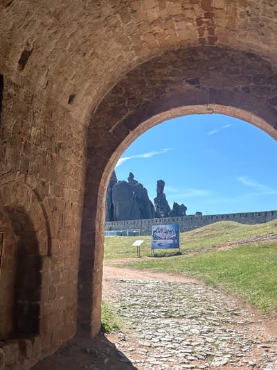 The image depicts an arched stone passageway looking out onto a landscape with distinctive rock formations. In the foreground, the rough-hewn stone walls and arched ceiling of the passageway are visible, casting shadows. Beyond the archway, a paved path leads towards a grassy slope. In the middle ground, a stone wall stretches horizontally, topped with battlements. Behind the wall, several tall, jagged rock formations dominate the skyline against a bright blue sky with sparse clouds. A sign with an informational map is placed to the left of the path, in front of the rock formations. The scene appears to be an outdoor historical or natural site, likely a fortress or protected area, given the architectural elements and the prominent rock formations. This view is consistent with the Belogradchik Rocks, a famous natural landmark in Belogradchik, Bulgaria. There are no people or animals visible in the image.