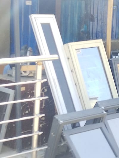 The image depicts an outdoor commercial or storage area in Darmanawa, Nigeria, primarily showcasing window and door frames. In the mid-ground, a tall, slender white frame with a dark grey or black insert stands upright, leaning against a blue corrugated metal structure. To its right, a beige-colored frame with a clear glass panel reflects light, showing a faint reflection of the surroundings. In the foreground, multiple grey-colored frames with lighter, possibly opaque, inserts are stacked horizontally. A silver-colored metal railing with four horizontal bars partially obscures the view in the lower left and center. In the blurry background, behind the frames and railing, machinery or tools are vaguely visible, along with a partially obscured individual in a light-colored shirt or garment, positioned to the left of the prominent white frame. The overall lighting suggests daylight.