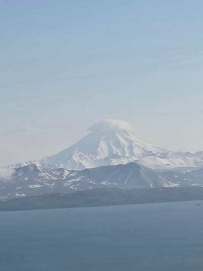 The image shows a distant, snow-capped volcano rising above a mountain range and a body of water. The sky is a pale blue with some hazy clouds. The foreground features a vast expanse of calm, blue water, with a small, dark ship visible on the right side. The middle ground is dominated by the jagged peaks of the snow-covered mountains. The volcano, the largest and most prominent feature, is in the background and is partially obscured by a swirling cloud near its summit. The scene is serene and majestic, conveying a sense of natural grandeur. The location is identified as being near the city of Krutoberegovy, Russia, which is known for its proximity to volcanic landscapes.