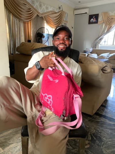 A man with a beard and baseball cap smiles, holding a torn pink "Hello Kitty" backpack. He wears an Apple watch and a white earbud. He is dressed in a cream-colored outfit and sits in a chair. A beige couch is behind him. Heavy beige drapes frame a window. An air conditioning unit is visible near the ceiling. A framed photo hangs on the wall. A fan is visible behind the man.