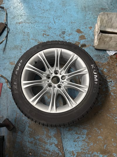A silver multi-spoke alloy wheel with a Kumho tire is resting on a blue, worn concrete floor. The tire has white lettering that reads "KUMHO" prominently on its sidewall, along with other technical specifications such as "245/45R18 100V XL" and "Asymmetric Snow Studless". The floor shows signs of wear and tear, with patches of lighter blue paint and darker, rougher concrete visible. In the upper left corner, a portion of a white object, possibly a container or equipment, is visible. There are also some loose wires and a small tool lying on the floor near the wheel. The scene appears to be in a workshop or garage setting. The lighting suggests it could be indoors during the daytime.