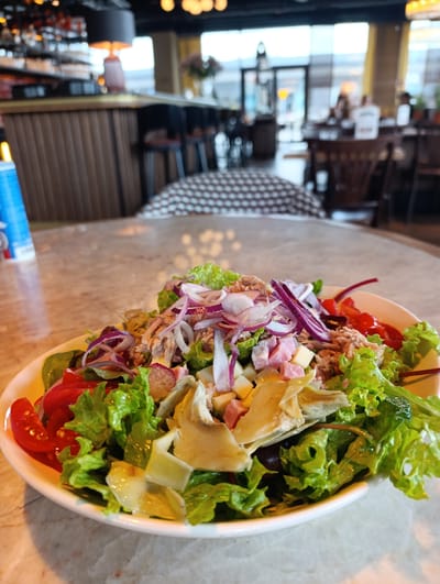 A close-up, eye-level shot captures a vibrant tuna salad served in a white bowl on a marble-patterned table. The salad is generously piled with crisp green lettuce, sliced red tomatoes, thinly sliced red onions, chunks of cheese, cubes of ham, and flaky tuna. The background is a blur of a restaurant interior, suggesting a casual dining atmosphere. The soft lighting highlights the fresh ingredients of the salad. The overall impression is one of a delicious and healthy meal presented in a modern establishment. Given the detail provided by the user, the location is Mysiadło, Poland.