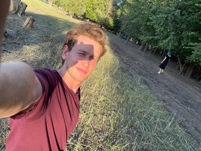 A young man with light brown hair takes a selfie outdoors. He is wearing a maroon t-shirt and is winking at the camera. In the background, a person with blue hair wearing a black dress is visible. The scene appears to be in a wooded area with dry grass and dirt paths. There is a light blue vehicle parked in the distance. The lighting suggests it is late afternoon or early evening. The location is identified as Rassvet, Kyrgyzstan.
