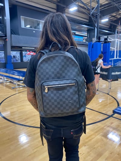 A person wearing a black t-shirt and dark jeans stands with their back to the camera on a polished wooden basketball court. They are carrying a checkered backpack. The person has long, dark hair and tattoos on their visible arm. In the background, another person is visible near a table with "molten" written on it. Bleachers and athletic equipment are also present in the indoor sports facility, which is located in Virginia Beach, United States of America.