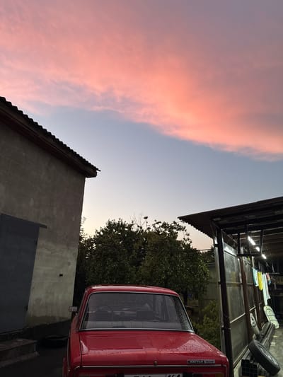 A red sedan is parked in front of a weathered building with a large gray garage door. To the right of the building is a dense green tree. Further to the right, a covered structure with lighting fixtures and clothes hanging on a line is visible. The sky above is a soft gradient of pale blue with streaks of pinkish-orange clouds, indicating either sunrise or sunset. The car appears to have water droplets on its surface. The location is identified as Novoshakhtinsk, Russia.