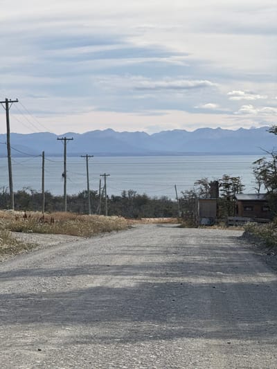 The image displays a rural landscape in Tolhuin, Argentina. A light grey gravel road, exhibiting tire tracks and minor undulations, occupies the foreground and recedes into the midground, curving slightly towards the right. On the left side of the road, a strip of dry, light-colored vegetation with visible white flowering or seeding heads borders the path. A sequence of five prominent wooden utility poles, fitted with overhead power lines, extends diagonally from the mid-left towards the background, paralleling the road. Beyond these poles and a line of sparse, possibly deciduous, trees or bushes, a vast body of calm, light blue-grey water, likely a lake or bay, stretches across the midground. In the far background, a horizontal range of blue-grey mountains with rugged peaks is visible, silhouetted against the sky. On the right side of the road in the midground, two structures are present: a small, weathered, light-colored wooden shack or shed with a dark chimney protruding from its roof, and behind it, a rustic, darker-toned wooden cabin or house. Scattered, sparse trees with minimal foliage surround these structures. The sky above is partially cloudy, featuring a mix of light blue and white-grey cloud formations. No individuals, actions, or interactions are depicted within the scene.