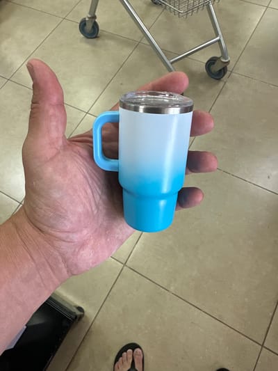 A close-up shot shows a person's hand holding a blue and white tumbler with a handle. The tumbler has a metallic silver lid. The background consists of beige tile flooring and the partial view of a metal shopping cart. A pair of black sandals is visible in the lower right corner of the image, suggesting a casual setting. The lighting indicates it's daytime, possibly indoors in a brightly lit area like a store or market. The focus is on the tumbler and the hand holding it, with no text visible in the frame. The overall impression is of a simple, everyday moment.