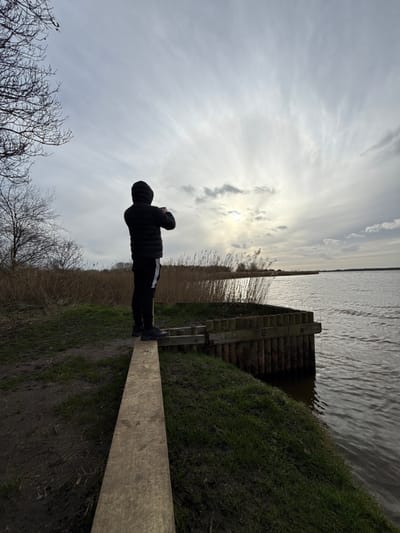 A lone individual stands balanced on a weathered wooden plank extending from a grassy embankment towards a body of water. The person, viewed from behind, wears a dark hooded puffer jacket, dark trousers with a white vertical stripe on the outer leg, and dark athletic shoes. They are holding an object, likely a mobile phone, with both hands, raised to capture an image or video. Adjacent to the plank and to the right, a wooden piling structure forms part of the bank's edge. The expansive body of water exhibits surface ripples and reflects a bright, overcast sky. The distant banks are lined with dense, tall, light-brown reeds. Bare deciduous tree branches are visible on the far left. The sky is predominantly overcast with wispy cloud formations, but the sun is visible breaking through the clouds near the horizon, creating a bright glare on the water. This environment is characteristic of the waterways and wetlands found within the Norfolk Broads, a national park area that extends into the North Norfolk district, United Kingdom.