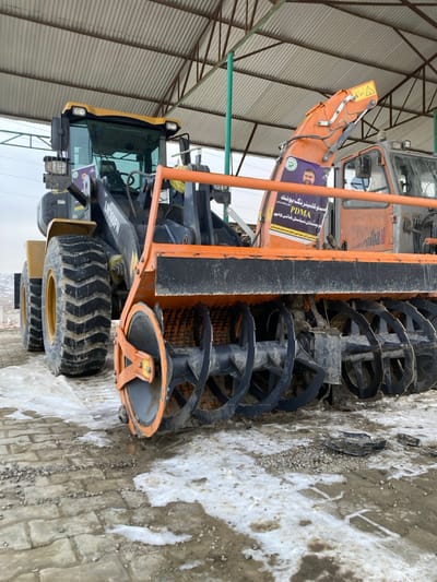 The scene depicts heavy machinery parked under an open-sided metal shelter in Kan Mehtarzai, Pakistan. In the foreground and slightly to the right, an orange snow blower attachment with prominent, dark-colored helical augers is visible, exhibiting dirt and remnants of snow. A purple and white banner, featuring a man's portrait and text (including "PDMA" and script identifiable as Urdu/Pashto), is affixed to the snow blower's discharge chute. To the left, a yellow and black wheel loader, identified by "LW300FN" on its side, is partially visible with large, robust tires. Behind the snow blower, a portion of another orange-toned heavy machine with "alba" visible on its side can be seen. The ground beneath the machinery is paved with light-colored interlocking bricks, partially covered with patches of melting snow, ice, and scattered gravel. Overhead, a corrugated metal roof supported by green structural beams provides cover. The distant background shows indistinct hills or structures under a pale, overcast sky. No individuals are present in the image.