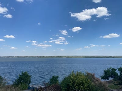 A wide-angle, landscape photograph captures a serene daytime view of a large body of water, likely the Dnieper River, from a slightly elevated perspective. The sky above is a vibrant blue, dotted with scattered white cumulus clouds. The water, a deep blue, exhibits gentle ripples, suggesting a light breeze. In the distance, a continuous line of low-rise buildings, characteristic of a town or village, stretches across the horizon, bordering the far bank of the river. The foreground features an embankment covered in dry grasses and various shrubs and trees, with some of the foliage appearing denser and greener than others. A few pieces of discarded building materials, possibly concrete slabs, are visible on the right side of the foreground, near the embankment. The overall scene evokes a peaceful, natural setting with a human settlement in the background.