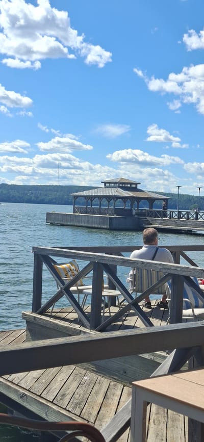 This is a sideways photograph taken on a sunny day with a bright blue sky dotted with fluffy white clouds. The scene is a wooden pier or boardwalk on a body of water. In the foreground, a man wearing a light-colored shirt and dark shorts is seated at a table, with his back to the camera. He has a dark strap across his shoulder. Several white chairs are arranged around tables, suggesting an outdoor seating area, possibly a cafe or restaurant. A yellow and white striped bag with text is visible near the base of the wooden structure.

Further in the distance, across the water, is a long, multi-story building with a distinctive roof structure. It appears to be built over the water, supported by stilts or pilings, and has large windows along its facade. The background shows a treed shoreline and more of the water. The overall impression is a serene outdoor setting, likely a recreational area or a waterfront dining establishment, on a pleasant day. The time of day appears to be daytime, with bright sunlight casting shadows. There is some visible text on the bag in the foreground, which appears to say "CARLTON BEACH".