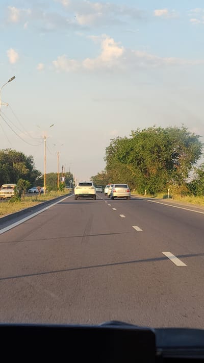 The image captures a view from a vehicle on a highway, with Tsiatsan, Armenia, suggested as the location. The highway is paved and has lane markings. Several cars are visible on the road, traveling in the same direction. On the sides of the highway, there are trees, vegetation, and utility poles with streetlights. The sky is partly cloudy. The image appears to have been taken during the daytime, possibly late afternoon or early evening, given the lighting.