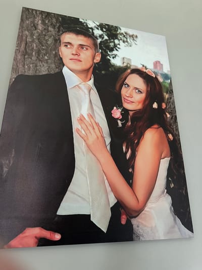 This is a painting of a newlywed couple posed for a portrait. The man is on the left, wearing a black suit jacket and a white dress shirt with a white tie. He is looking to his left, with a serious expression. The woman is on the right, wearing a white wedding dress and a floral hairpiece. She is looking at the viewer with a slight smile. Her hand is resting on the man's chest. The background appears to be a park-like setting with trees and buildings in the distance. The style of the painting is realistic with a soft, painterly quality.
There is no information available in the image or the crops to suggest that the location is the city of Konstantinovo, Russia.