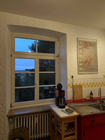 The image depicts a domestic kitchen scene at dusk, with a view of a rural landscape through a large window. The kitchen features white, textured walls and a white-framed window with multiple panes. Outside the window, the sky is a deep blue, suggesting twilight, and trees and fields are visible. Inside, a red and chrome coffee maker sits on a wooden cart next to the window. To the right, a red countertop with a stainless steel sink and faucet is visible. Above the countertop, a tiled backsplash with yellow and white squares is present. A framed poster with an anatomical drawing and text in German hangs on the wall. A white radiator is situated beneath the window, with a brown paper bag and other items placed in front of it. The overall impression is of a functional, lived-in kitchen in a rural or semi-rural setting.