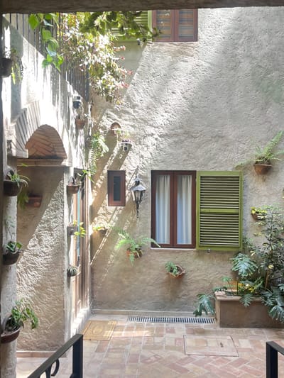 The image captures an enclosed outdoor courtyard or patio within a building complex in Santa Ana Portales, Mexico. The walls are constructed of light beige, textured stucco, displaying prominent shadows cast by direct sunlight from the upper left. The ground is paved with reddish-brown bricks arranged in a herringbone pattern, featuring two linear metal drainage grates.

On the left, a brick archway provides access to an adjacent area, and directly to its right, a recessed tall, narrow wooden door with multiple panes is present, with a light-colored patterned doormat positioned before it. A small, white dome-shaped security camera is mounted on the stucco wall above the door.

The central wall features a small, vertically oriented brown wooden window, below which an ornate dark metal lantern is mounted. To the right of the lantern, a larger double-paned brown wooden window with white interior curtains is visible, flanked by a single open light green louvered shutter on its right. In the upper right corner, a brown wooden window with closed wooden shutters is partially visible.

The environment is verdant with various plants: several small ferns and leafy plants are situated in terracotta-colored hanging pots affixed to the walls, green foliage cascades from an upper railing on the left, and larger green plants are contained within a rectangular ground-level planter on the right. A dark metal railing is present in the immediate foreground, extending from the bottom left. The scene is devoid of people.