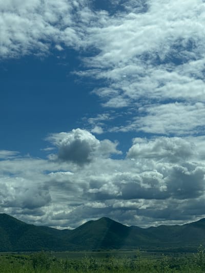 The image displays a wide, panoramic view of a mountainous landscape under a dynamic sky filled with white and gray clouds. The foreground is dominated by lush green vegetation and trees, suggesting a natural, undeveloped area. Beyond the foreground, rolling hills covered in dense, dark green foliage rise, forming a series of mountain ranges that stretch across the horizon. The sky is a vibrant blue, with scattered clouds ranging from fluffy white cumulus to darker, more ominous gray formations, indicating variable weather conditions. The overall scene depicts a natural environment, possibly a rural or wilderness area, characterized by its extensive greenery and cloud-filled sky. No people, man-made structures, or specific landmarks that would pinpoint the location to Теплоозёрск, Russia, are visible in the image.